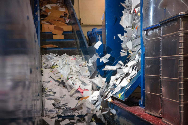 Papierflut auf Förderband, bunte und weiße Blätter, Kartons, Metallbehälter, Recyclinganlage.